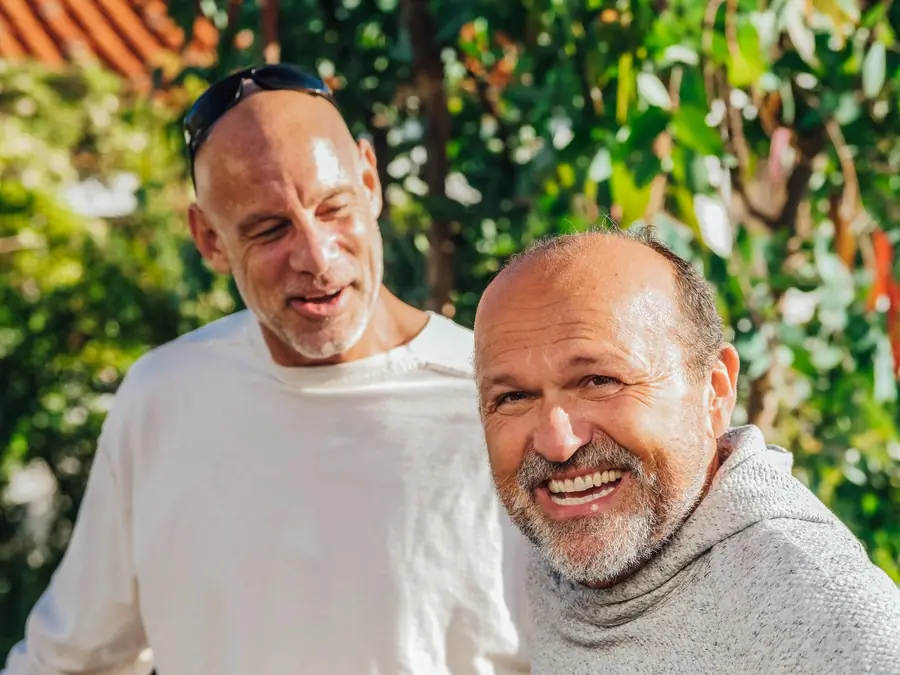 Two older men sitting close together outdoors, both smiling